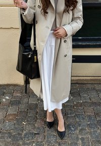 Trench coat beige de doble botonadura, falda midi blanca, tacones negros tipo slingback y un bolso de hombro de cuero negro. Fondo de adoquines texturizados.