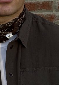 Sciarpa paisley marrone indossata con una camicia scura a bottoni sopra una maglietta bianca. Texture visibile e dettagli delle cuciture sulla camicia.