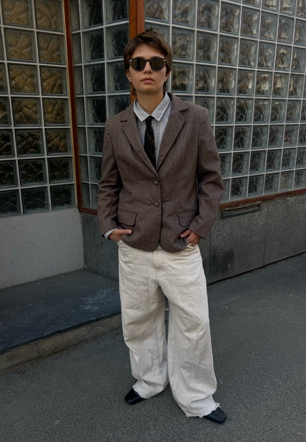 Personne portant des lunettes de soleil, un blazer marron, une chemise à carreaux avec une cravate noire, un pantalon blanc large, debout les mains dans les poches près d’un mur en blocs de verre.