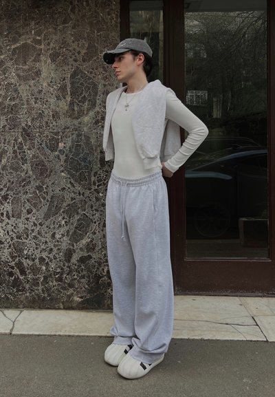 Bodysuit blanco de manga larga, chaleco sin mangas gris, pantalones de chándal gris claro, zapatillas blancas de gran tamaño con acentos negros, todo con un fondo de mármol.