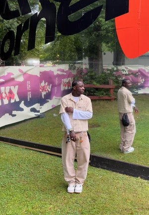 Un fondo de espejo reflectante con patrones de camuflaje vibrantes en púrpura y rosa. Una persona con una camisa de cuadros beige y pantalones claros está de pie frente a él.