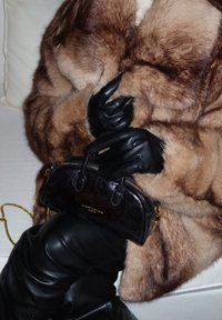 Borsa nera in pelle con dettagli dorati. Cappotto di pelliccia in tonalità di marrone. Guanti lunghi neri. Pantaloni neri in pelle, seduta su un divano chiaro.