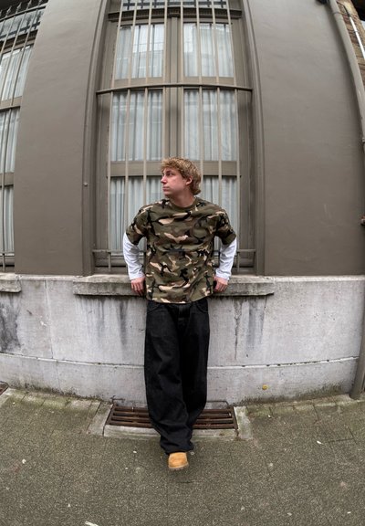 Camisa de manga corta con patrón de camuflaje, superpuesta a una camisa de manga larga blanca, combinada con pantalones vaqueros holgados negros y zapatos marrones, apoyado contra una pared.