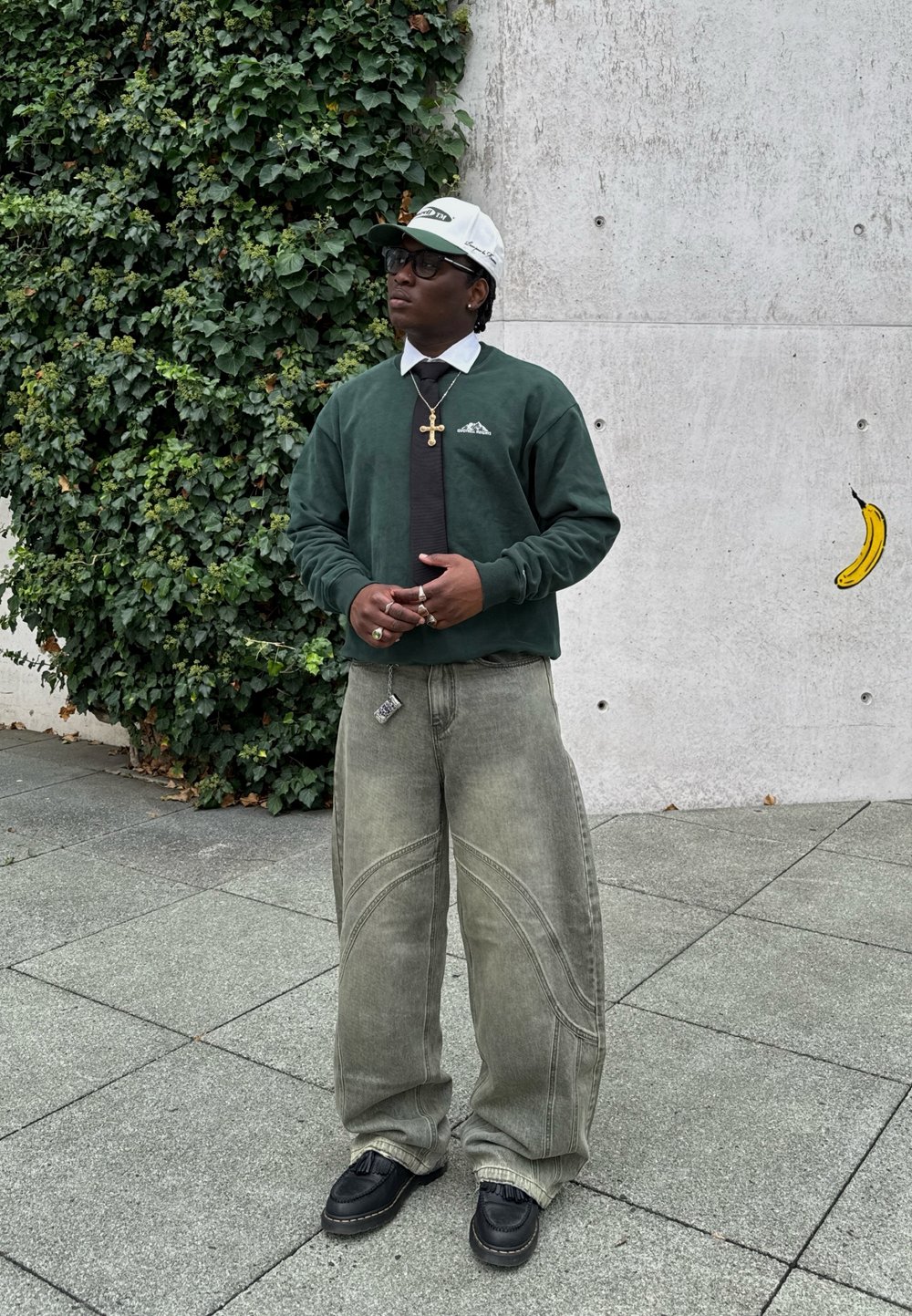 Green sweatshirt with logo, white shirt, black tie, grey wide-leg jeans with stitched patterns, and black shoes. The background features greenery and concrete.