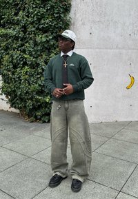 Green sweatshirt with logo, white shirt, black tie, grey wide-leg jeans with stitched patterns, and black shoes. The background features greenery and concrete.