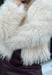 Puhast bela umetna krznena jakna z dolgim, valovitim teksturiranim vzorcem in volumnastimi rokavi, nošena preko oprijete bordo usnjene krila.