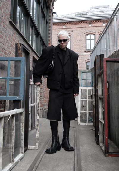 Persona con pelo corto rubio que lleva una chaqueta negra de gran tamaño, pantalones cortos, botas y gafas de sol, de pie entre viejos marcos de ventanas en un callejón urbano.