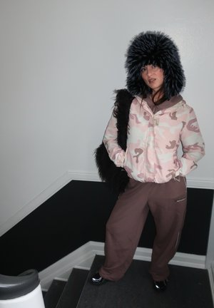 Chaqueta de plumas de camuflaje rosa con capucha de piel sintética, combinada con pantalones de chándal marrones y calzado negro brillante. Bolso de hombro negro texturizado.
