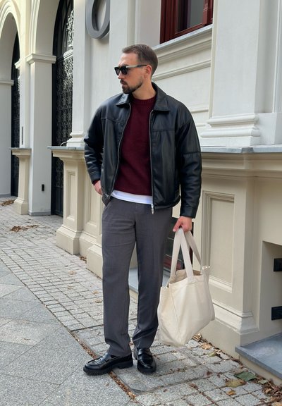 Chaqueta de cuero negra sobre un suéter burdeos, pantalones grises, zapatos negros, sosteniendo una bolsa de mano beige. Texturas suaves, ajuste moderno, entorno urbano.