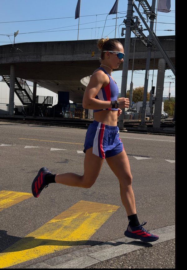 Femme athlétique en tenue de course bleue et rouge jogge dans la rue près d'un passage inférieur, portant des lunettes de soleil et des chaussures de course noires avec des rayures rouges.