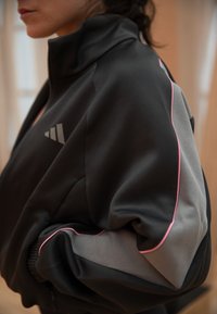 Κοντινό πλάνο ατόμου που φοράει μαύρο και γκρι μπουφάν Adidas με ροζ λεπτομέρειες, στέκεται εσωτερικά μπροστά σε θολό φόντο.