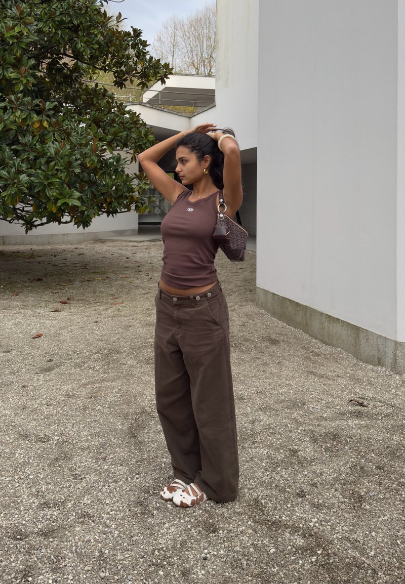 Woman with dark hair in brown sleeveless top and wide brown pants adjusts hair outdoors near modern white building and green tree.