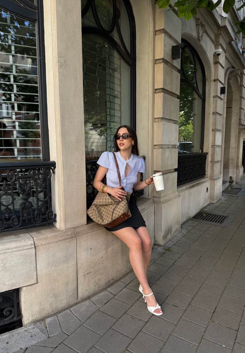 Femme avec des lunettes de soleil assise sur un rebord en pierre à l'extérieur d'un bâtiment, tenant une tasse blanche, portant un chemisier bleu, une jupe noire, des talons blancs et portant un sac à motifs.