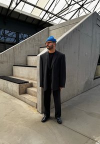 Μαύρο υπερμεγέθες σακάκι και παντελόνι, μαύρο πουκάμισο, μπλε πλεκτό σκουφί, μαύρα παπούτσια, στέκετ�αι κοντά σε σκυρόδεμα σκαλιά σε έναν σύγχρονο αρχιτεκτονικό χώρο.