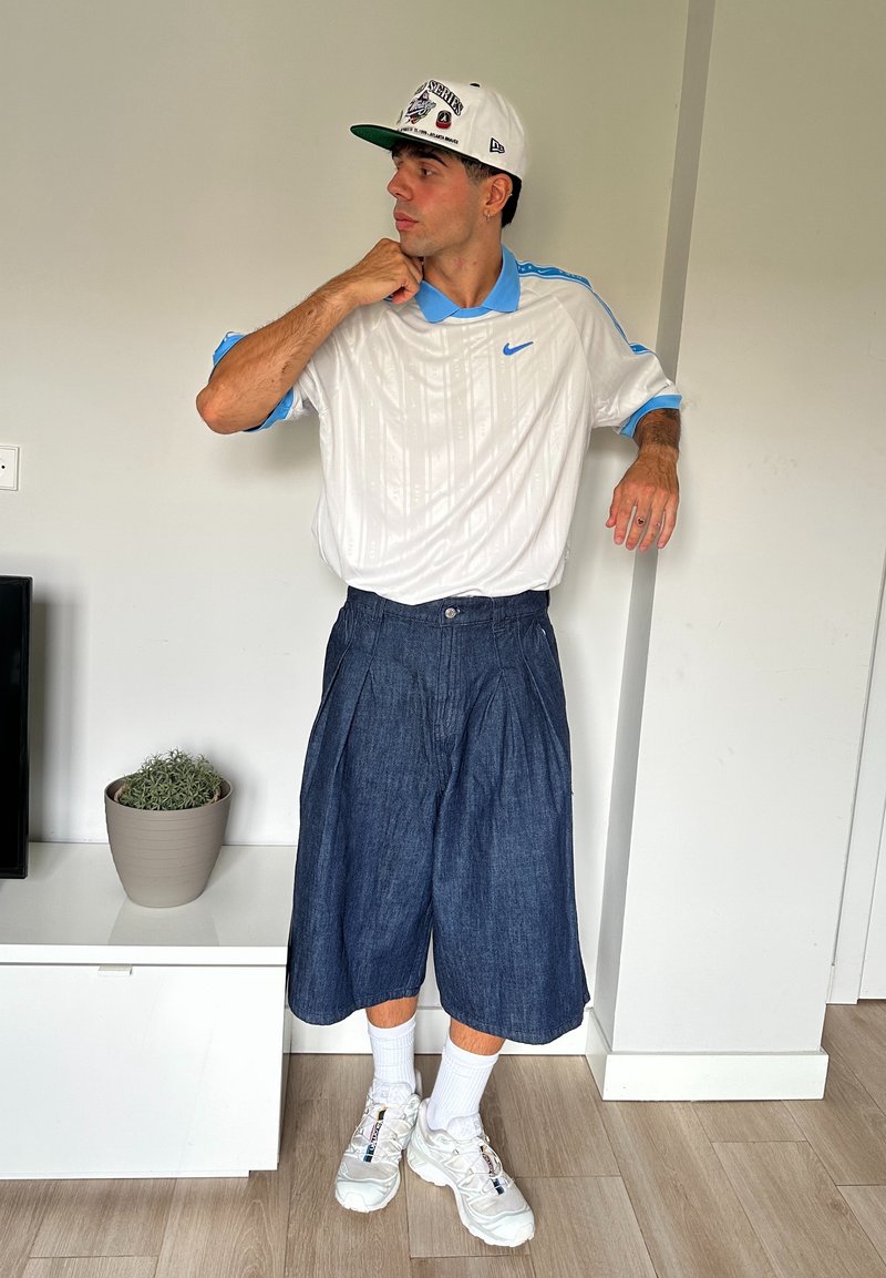 Polo shirt blanc Nike avec des accents bleu clair, un short en denim foncé plissé, des chaussettes blanches et des baskets blanches. L'arrière-plan comprend une plante en pot.
