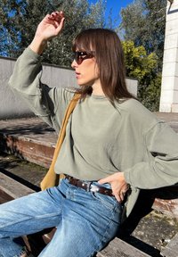 Top de mangas longas em verde oliva com nervuras, combinado com jeans azul claro, cinto castanho e mala de ombro castanha; óculos de sol usados.