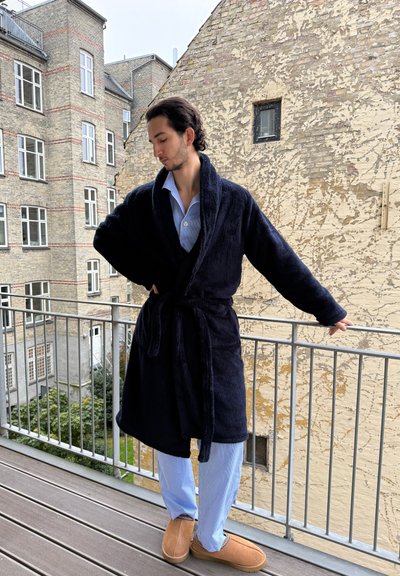 Bata de felpa navy con cuello de chal y cinturón, llevada sobre pijamas a rayas azul claro. Un par de zapatos marrones de tipo deslizable completan el atuendo. Entorno en un balcón exterior.