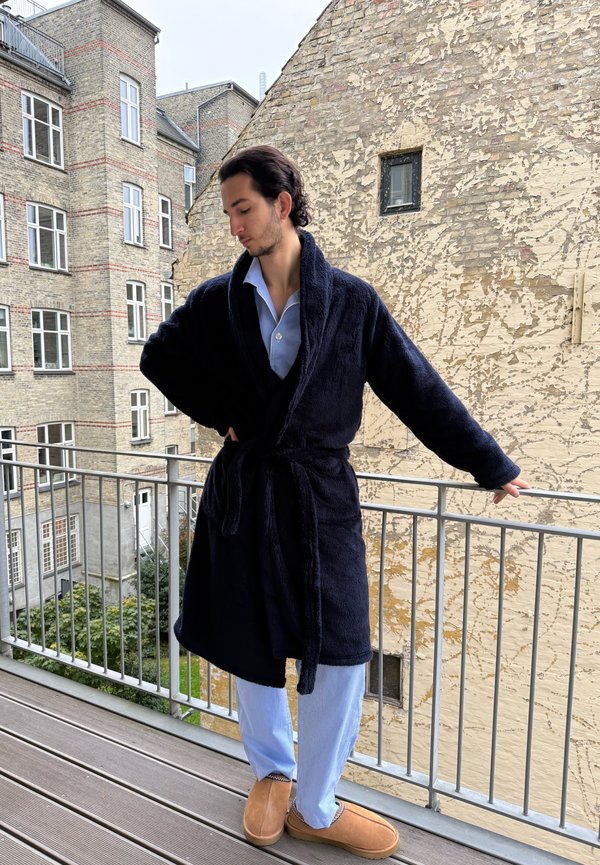 Granatowy pluszowy szlafrok z kołnierzem szalowym i paskiem, noszony na jasnoniebieskich, w pionowe paski piżamach. Brązowe buty wsuwane dopełniają strój. Ustawienie na zewnątrz, na balkonie.