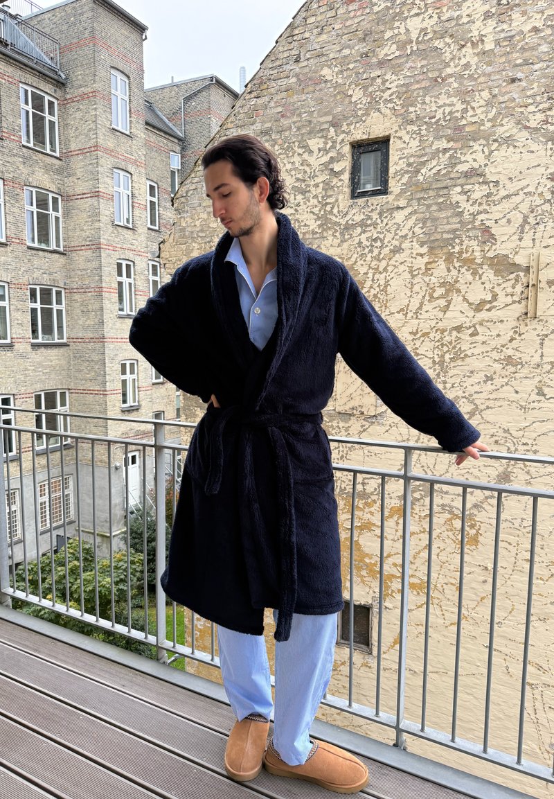 Navy plush robe with shawl collar and tie belt, worn over light blue striped pajamas. Brown slip-on shoes complete the outfit. Outdoor balcony setting.