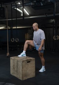 Caixa de pliometria em madeira com uma superfície lisa. Pessoa a subir, vestindo roupa de desporto de cor clara e sapatilhas com detalhes azuis.