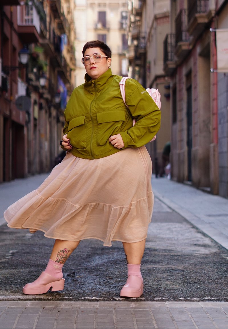 Persoon met een groene jas, een beige vloeiende rok, roze schoenen en sokken, en een roze rugzak, die zelfverzekerd staat op een smalle stadstraat.