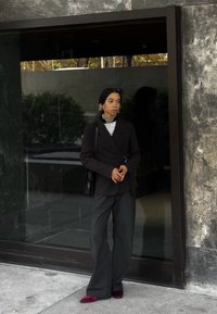 Melna pielāgota žakete ar ietīšanas dizainu, saskaņota ar plašām pelēkām biksēm un bordo kurpēm. Iekļauj šķērsotu apkakli un lielas auskars.