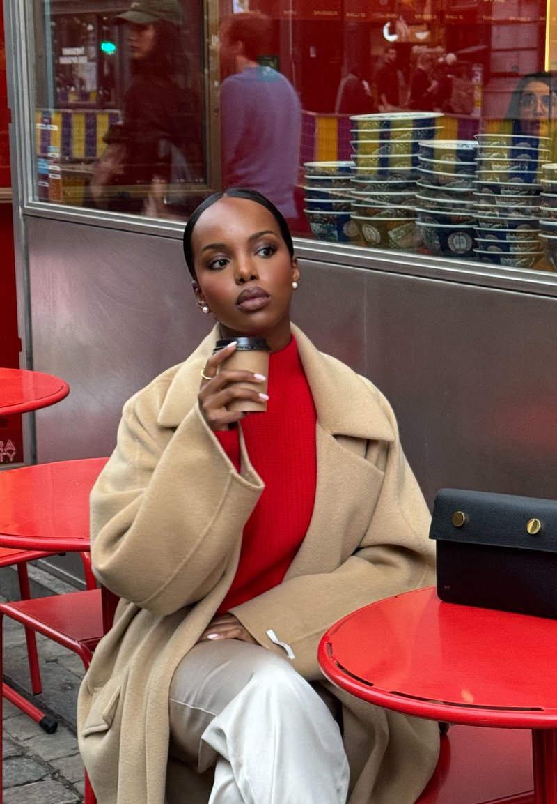 Cappotto di lana beige, maglione rosso, pantaloni crema. Tiene in mano una tazza di caffè. Borsa nera con dettagli dorati su un tavolo rotondo rosso, sfondo urbano.