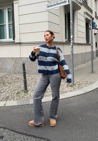 Gestreifter Cardigan in Marineblau und Grau, knöpfbar, kombiniert mit grauen Schlagjeans und beigefarbener Schuhen. Hält eine Tasse Kaffee und steht im Freien.