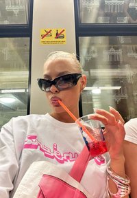 Άτομο που κρατά ένα διάφανο ποτήρι με κόκκινο ρόφημα και πορτοκαλί καλαμάκι. Φοράει λευκό πουκάμισο με ροζ σχέδια και ροζ τσάντα σώματος.