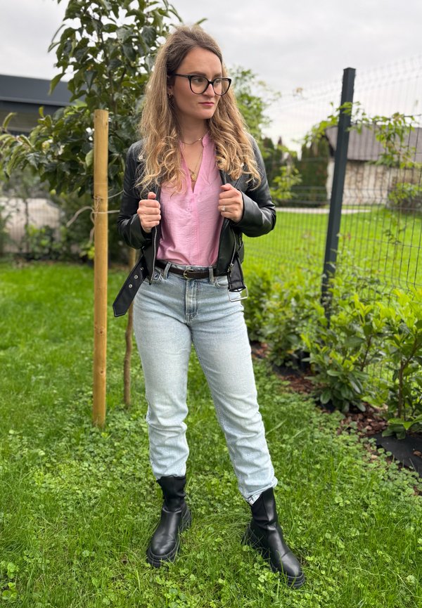 Black leather jacket over a pink blouse, teamed with light blue jeans and black ankle boots. Green grass and shrubs in the background.