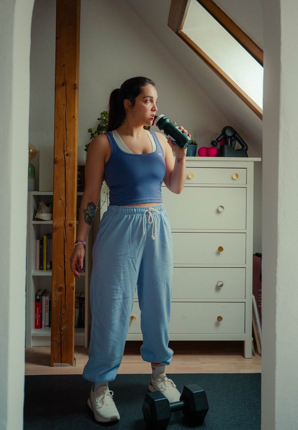 Vrouw in sportkleding drinkt uit een waterfles, staat op een mat met een dumbbell in een lichte zolderkamer.