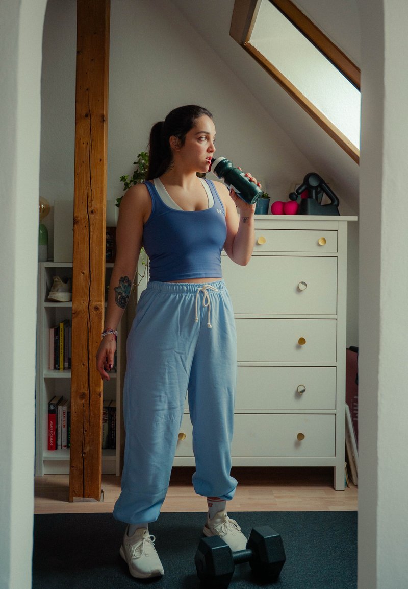 Vrouw in sportkleding die uit een waterfles drinkt, staand op een mat met een halter in een lichte zolderkamer.