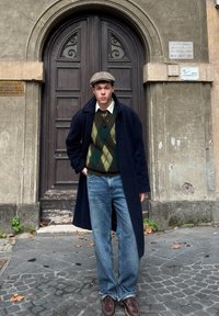 Manteau navy, pull argyle vert et marron, chemise claire, jeans bleus, chaussures brunes, debout devant une porte en bois avec des détails décoratifs.