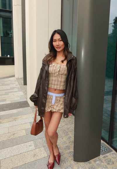 Chaqueta de cuero marrón sobre un top de cuadros corto y pantalones cortos a juego. Unos zapatos planos rojos de punta y un bolso marrón completan el conjunto. Texturas suaves.