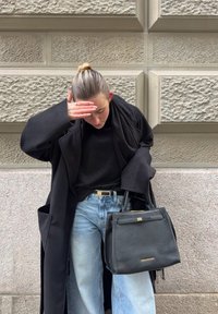 Γυναίκα με μαύρο παλτό και κασκόλ που καλύπτει τα μάτια της με το χέρι, κρατώντας μαύρη τσάντα, στέκεται μπροστά σε φόντο με υφή από πέτρινο τοίχο.