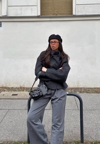 Černá bunda z vlněné směsi s vysokým límcem a knoflíkovými detaily, v kombinaci se šedými širokými kalhotami; černá kožená kabelka přes rameno a baret.