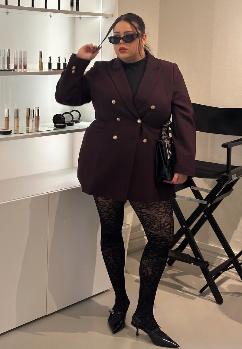 Burgundy double-breasted blazer with gold buttons, paired with black lace tights and pointy black heels; accessories include sunglasses and a handbag.