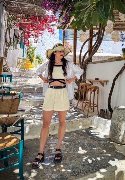 Camisa blanca, top corto negro y pantalones cortos color crema. Chanclas negras con suelas gruesas. Entorno al aire libre con suelo de piedra, vegetación y flores rosadas.