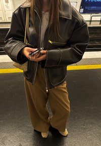 Barna bőrkabát cipzárral az elején és széles ujjakkal, világosbarna, széles szárú nadrággal és bézs lábbelivel párosítva, kezében okostelefont tartva.