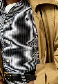 Beige trenchcoat over een zwart-wit gestreept shirt met een klein logo, gecombineerd met een zwarte riem met gouden hardware.