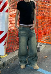 Pantaloni in denim a gamba larga di colore azzurro chiaro con una texture sbiadita, indossati con una maglietta nera e stivali beige, davanti a uno sfondo di recinzione di sicurezza arancione.