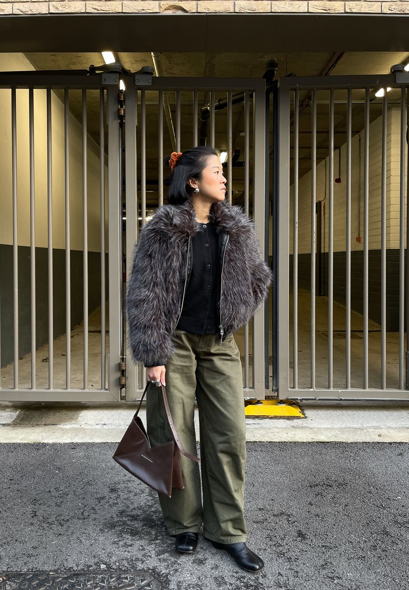 Femme debout à l'extérieur devant des portes métalliques, portant une veste en fourrure grise, un pantalon olive, des chaussures noires et tenant un sac à main brun.