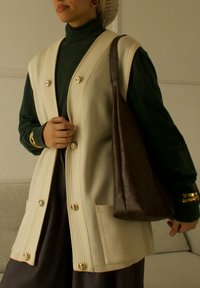 Personne portant un gilet beige boutonné sur un col roulé vert foncé, portant un grand sac à bandoulière en cuir marron, avec des bracelets et des boucles d'oreilles en or.