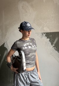 T-shirt court camouflage gris avec texte blanc, pantalon gris à cordon de serrage, et sac à fini métallique avec un design structuré et une bandoulière.