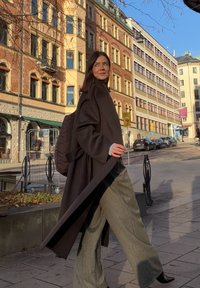 Long manteau brun foncé porté sur un pantalon gris à fines rayures. Le manteau présente un col haut et des manches larges. Un sac à dos matelassé et sombre est porté sur le côté.