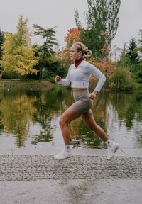 Lätt, långärmad vit topp, röd scarf, beige höga shorts och vita sneakers. Springer vid en reflekterande vattenspegel med träd.