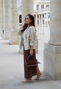 Giacca grande beige, abito maxi a pois marrone, décolleté rosa con tacco a rocchetto e una borsa marrone con un accento bianco. Pilastri di pietra sullo sfondo.