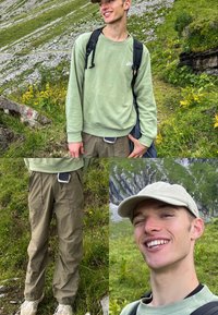Mladý muž v světle zelené mikině, béžové kšiltovce a turistických kalhotách se usmívá, zatímco stojí na travnaté horské stezce s kamenitými svahy.