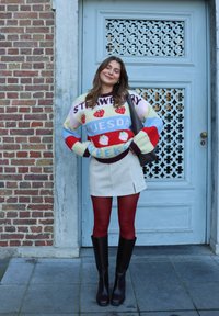 Jeune femme portant un pull coloré à thème fraise, une jupe blanche, des collants rouges et des bottes noires, se tenant devant une porte décorative bleue.