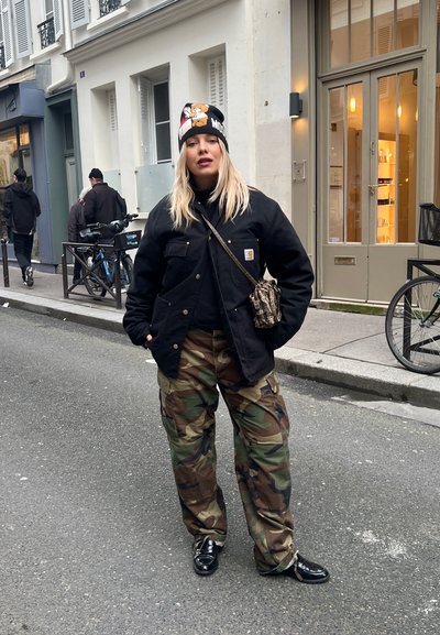 Chaqueta negra con un parche, jersey de cuello alto negro, pantalones de camuflaje y zapatos negros brillantes. Lleva un gorro de lana con personajes de dibujos animados. Escena urbana en la calle.
